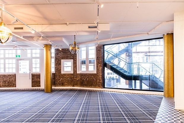 Bright open-plan commercial interior with exposed red-brick walls, gold cylindrical columns, large industrial windows, a glazed stair enclosure, decorative pendant lighting, and patterned carpet flooring. The space appears to be a lobby, office, or hospitality venue.