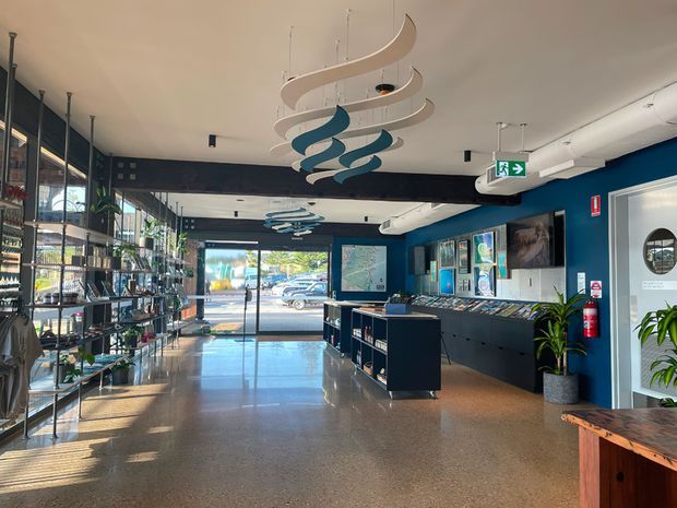 A bright contemporary commercial gallery or showroom interior with polished terrazzo flooring, deep blue walls, extensive black metal and glass display shelving, built-in display cabinetry, framed artwork, and a glazed street-facing entrance. Sculptural suspended white and blue ceiling elements create a prominent feature, while plants and a timber counter add warmth. An exit sign, fire extinguisher, and circular-windowed door are visible.