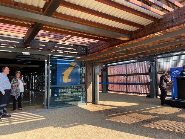 A covered entry or event space with exposed dark timber beams, corrugated translucent roofing, steel structural members, and patterned metal security screens. Glass doors open into a dark commercial interior with a prominent yellow logo, while a small presentation or service counter is visible on the right. Strong sunlight creates striped shadows across the textured floor.