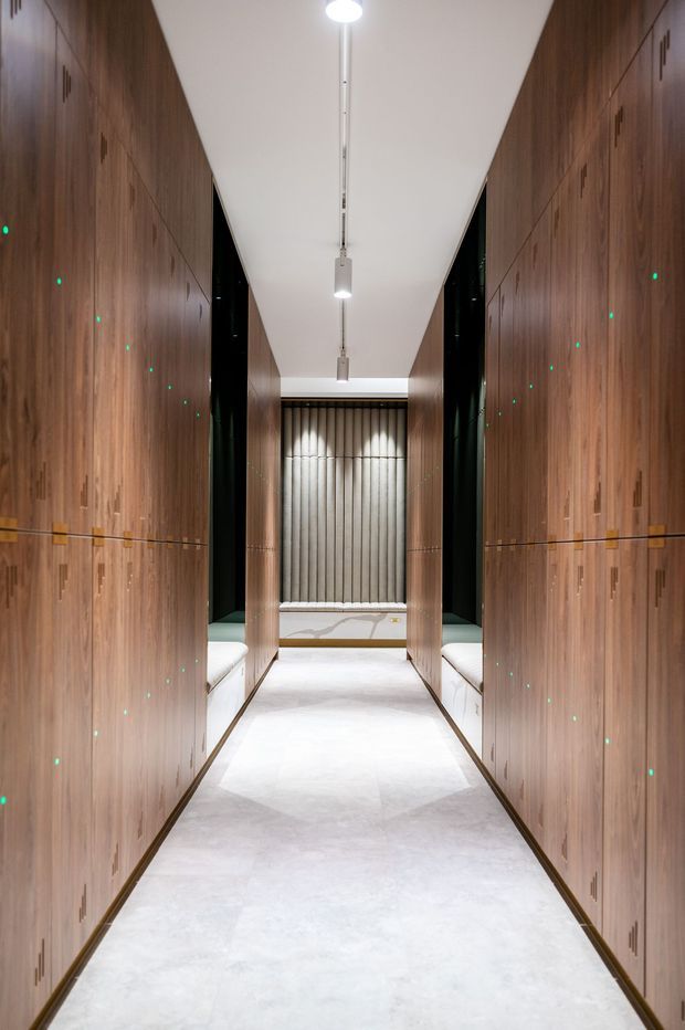 A narrow, symmetrical interior corridor lined on both sides with full-height dark timber cabinetry and integrated small handles. The pale polished floor and white ceiling create a bright, gallery-like passage, with slim suspended linear lights overhead. Dark glazed or reflective recesses and white marble-like benches appear at the far end, with a vertically ribbed feature wall beyond. Small green indicator lights punctuate the cabinetry.