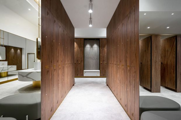 A refined contemporary dressing and bathroom area with full-height dark timber built-in cabinetry forming a central passage. The space has pale stone or concrete-look flooring, recessed ceiling lighting, a freestanding white basin and sculptural stone vanity visible to the left, and dark upholstered seating in the foreground. A tall narrow window with sheer curtains is visible at the end of the corridor.