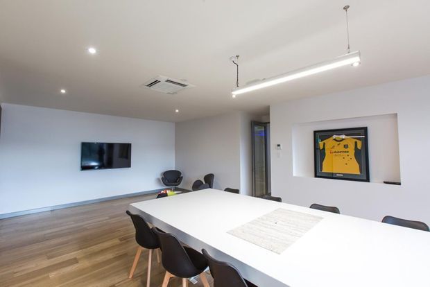 Bright contemporary dining and meeting area with a large white rectangular table, black molded chairs, timber flooring, white walls, linear suspended lighting, recessed ceiling lights, wall-mounted television, and a framed sports jersey displayed in a wall niche.