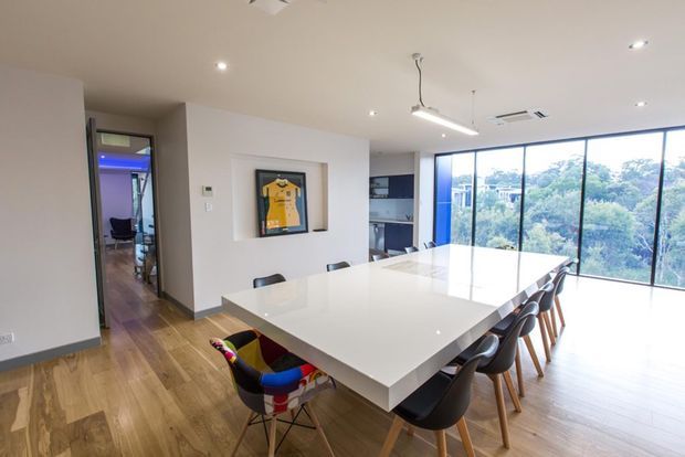 Bright contemporary open-plan dining area with a large white rectangular dining table and mixed modern chairs, set against warm timber flooring. Floor-to-ceiling glazing provides expansive views of leafy surroundings, while a compact kitchen is visible behind the dining space. Recessed ceiling lights and a linear pendant add clean modern detailing; a framed sports jersey provides a colourful accent.