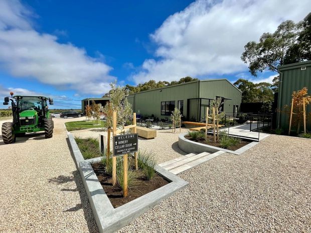 A landscaped gravel forecourt leading to a low-rise dark green corrugated-metal building complex. A sign welcomes visitors to a winery cellar door, with raised concrete-edged garden beds, young trees, ornamental grasses, paved paths, outdoor seating, and a tractor visible to the left. The scene is photographed outdoors in bright daylight with a blue sky and surrounding trees.