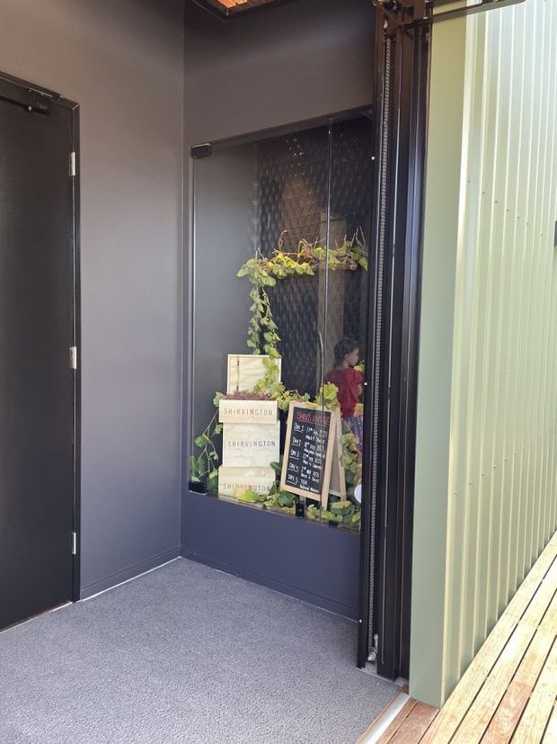 A compact, dark-toned entryway or transitional lobby with charcoal painted walls, a grey carpeted floor, and a black-framed textured-glass display window containing plants, stacked boxes, and a small chalkboard sign. A black door is visible on the left, while a large glazed or screened opening and pale corrugated wall panel appear on the right beside a timber decking strip. The space has an industrial, commercial character with warm natural accents from the plants and timber.