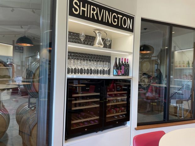 A built-in wine and glassware display in a refined hospitality or dining interior. The illuminated shelving is labelled “SHIRVINGTON” and holds wine glasses, decanters, bottles, and two under-counter wine refrigerators. Black-framed glazed partitions reveal a contemporary dining area with curved seating, pendant lighting, and reflective surfaces.