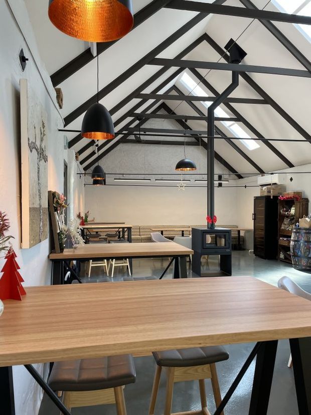 A spacious, barn-like dining and hospitality interior with long timber tables, timber-framed chairs, black steel table bases, exposed dark structural roof trusses, and multiple pendant lights. The white walls and vaulted ceiling create an airy setting, while a central black wood-burning stove, counter area, and display shelving add a rustic industrial character.