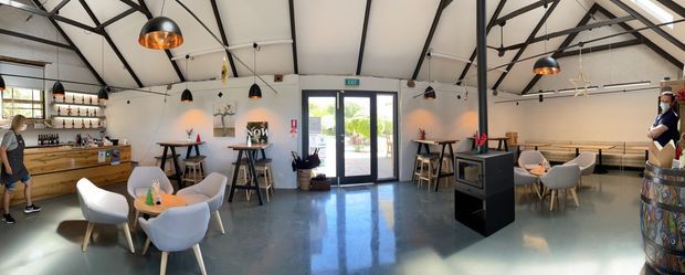 A bright, open-plan hospitality interior with polished concrete flooring, white walls, exposed black steel roof trusses, and pendant lighting. The space includes a timber service counter and shelving on the left, several small dining and lounge tables with upholstered chairs, a central freestanding display or service unit, and a long built-in timber bench along the right wall. Glazed double doors open to the exterior at the rear. The overall aesthetic is contemporary with industrial structural detailing.