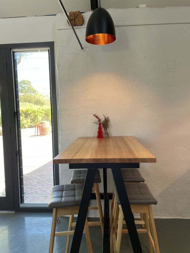 A compact dining area with a tall rectangular timber table and black A-frame metal supports, surrounded by upholstered bar stools with timber legs. A black pendant light with a warm copper-toned interior hangs above the table. The space has white textured walls, a polished grey concrete floor, and dark-framed full-height glazing opening to an outdoor paved area.