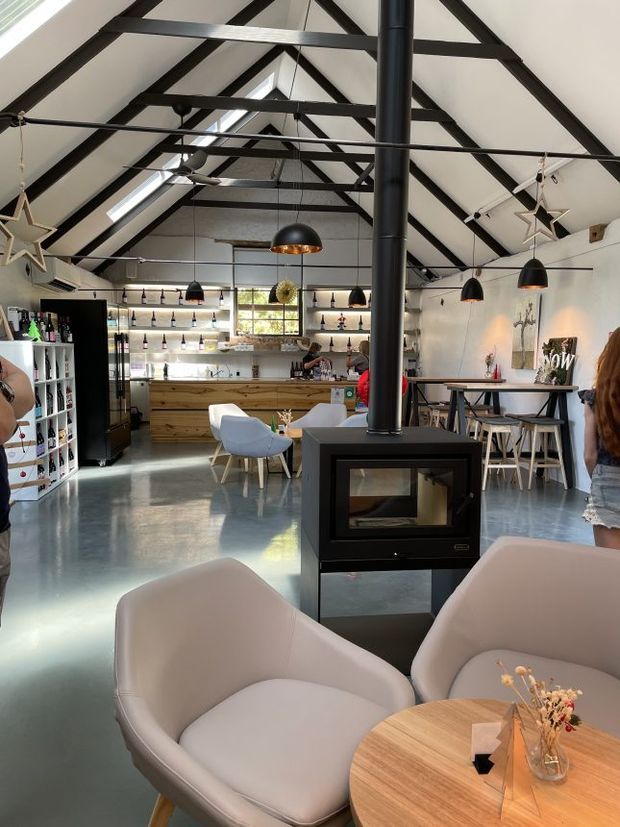 A spacious, high-ceilinged café or hospitality interior with a prominent exposed dark timber truss roof, white ceiling panels, skylights, and polished concrete flooring. The room includes a timber service counter and open shelving, pendant lighting, bar-height and dining seating, upholstered lounge chairs, and a freestanding black wood-burning stove with a flue extending to the roof. The atmosphere is warm, rustic-industrial, and inviting.
