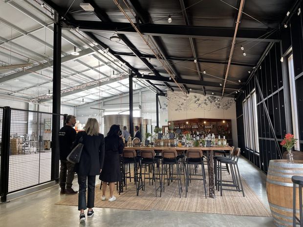 A spacious industrial-style commercial bar and dining area with a long timber bar counter, numerous high stools, bottle displays, pendant and track lighting, exposed services, black-painted ceiling, and large steel-framed glazed partitions. Visitors are present in the foreground, while a barrel table and bar furnishings add to the hospitality setting.