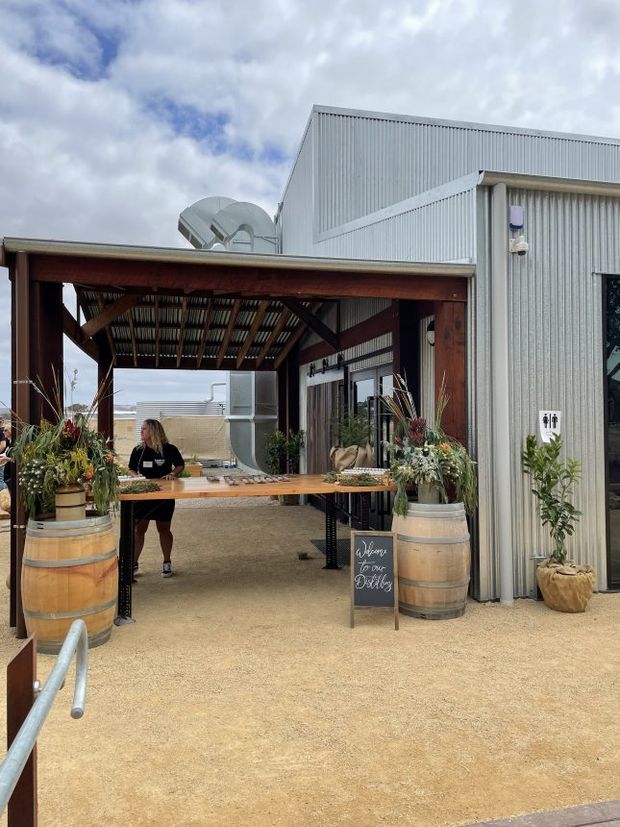 Exterior view of a corrugated metal-clad commercial or event building with a timber-framed covered entry canopy. The open-air entrance includes a long timber reception or serving table, floral arrangements in wooden barrels, a freestanding welcome sign, and accessible-looking paved ground. The scene appears to be an event or hospitality entrance, with industrial materials softened by timber and planting.