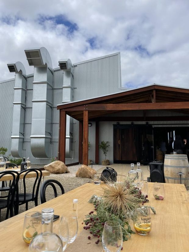 An outdoor dining and hospitality area beside a corrugated-metal industrial building. A large timber-framed covered structure shelters a dark entrance, while a long timber dining table is set with glassware, bottles, floral arrangements, and black bentwood-style chairs. Large exposed metal ductwork is visible on the building facade, with gravel landscaping, rocks, and a wooden barrel adding to the rustic industrial atmosphere.