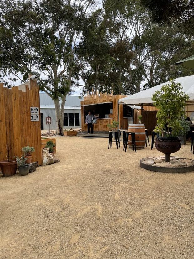 An outdoor hospitality courtyard set among mature eucalyptus trees, with a timber-clad bar kiosk, covered seating area, high stools, barrel-style tables, potted plants, gravel paving, and a raised circular planter. The setting has a rustic, relaxed garden character with timber fencing and light metal-roofed structures.