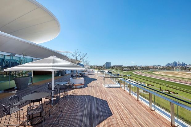 A wide outdoor terrace with timber decking, café-style tables and chairs, large umbrellas, planters, and glass balustrades overlooking a horse-racing track. The terrace is attached to a sleek contemporary building with sweeping curved roof forms, with a distant urban skyline beyond.