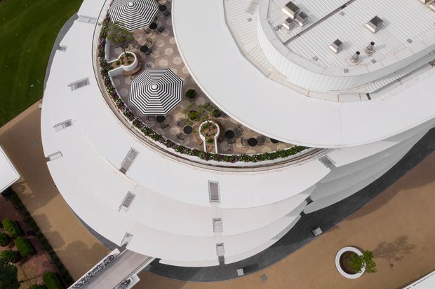 Aerial view of a large curved, layered white roof terrace or building complex. The central terrace contains patterned outdoor seating, striped circular umbrellas, planted garden beds, and paved walkways, with additional rooftop equipment and circular architectural forms visible around it.