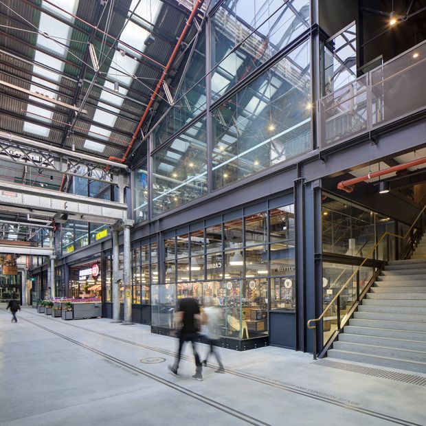 Interior view of a large adaptive-reuse style commercial arcade or market hall, featuring exposed steel framing, corrugated metal roofing, extensive glass shopfronts, overhead services, concrete flooring, and a broad stairway leading to an upper level. Retail and food outlets line the circulation space, with blurred pedestrians indicating an active public setting.