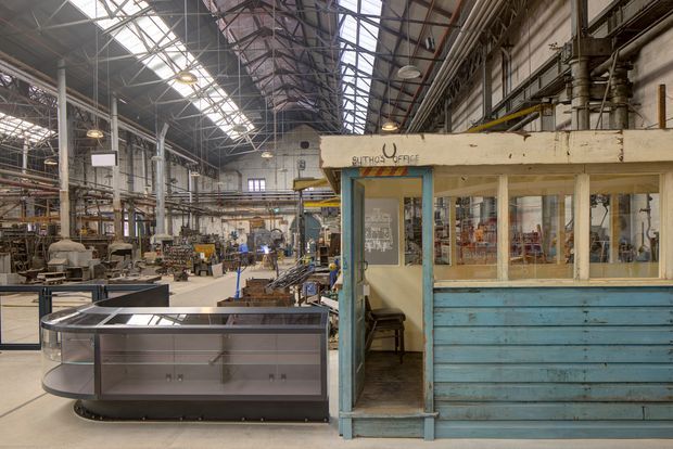 A large adaptive industrial interior resembling a historic factory or workshop, with exposed steel roof trusses, extensive overhead glazing, industrial machinery, workstations, and a small weathered office booth in the foreground. The booth features pale blue timber cladding, cream-painted framing, glazed panels, and an open doorway. A curved glass-fronted display or work counter sits in front, while the expansive factory floor extends into the background.