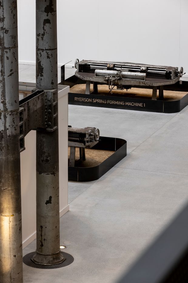 A contemporary museum or exhibition interior displaying historic industrial machinery, including a labelled Ryerson spring forming machine, on black-edged plinths. Weathered steel structural columns, white walls, and a smooth pale grey concrete floor create an industrial gallery setting with strong directional shadows.