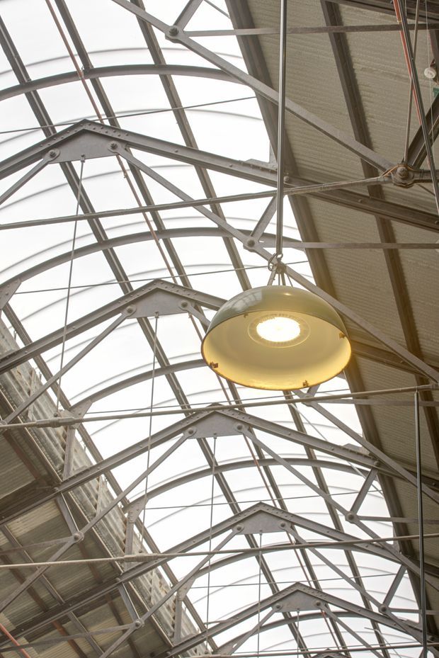 Upward-looking view of a large vaulted industrial roof structure, with repeating exposed steel trusses, translucent arched roof panels, corrugated metal sections, suspended cables, and a prominent circular pendant light. The image emphasises the dramatic structural framework and high ceiling of an industrial or commercial interior.