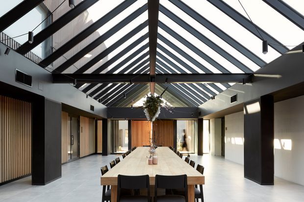 A dramatic, contemporary double-height dining hall with a very long timber dining table and dark upholstered chairs. The space is organised beneath a transparent pitched glass roof with exposed dark structural rafters and beams, creating a bright central atrium. Dark charcoal columns and framing contrast with pale walls and flooring, while timber slatted wall detailing, warm timber doors, pendant lights, and a central hanging plant add texture and warmth.