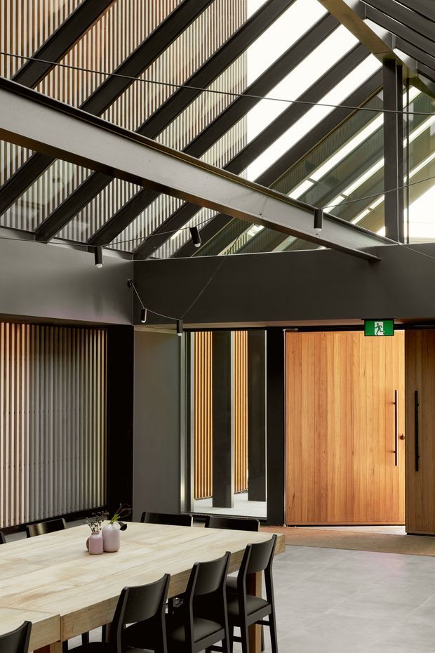 A contemporary communal dining or meeting space with a large light timber table and black chairs in the foreground. The room features dark exposed steel roof beams, a high glazed roof, vertical timber and metal screening, dark walls, suspended track-style lighting, and a tall timber pivot door with an emergency exit sign. The architecture has an industrial character softened by warm timber detailing and abundant daylight.