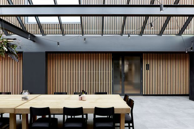 A contemporary dining area arranged around a long light timber dining table with dark chairs. The space features dark charcoal walls, extensive vertical timber battens, large pale grey floor tiles, and a glazed overhead roof structure with exposed dark beams and linear pendant lighting. A central dark-framed doorway and slatted screening define the rear wall, creating a refined indoor-outdoor architectural feel.