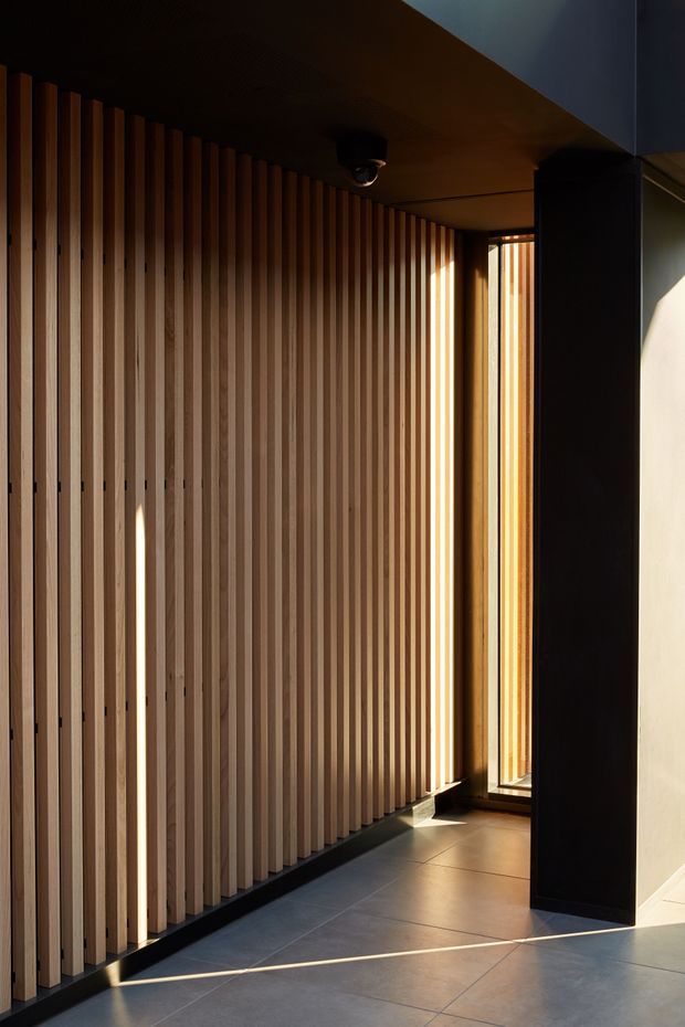 A dramatic modern interior corridor or entry space featuring a full-height vertical timber batten screen along the left wall, dark ceiling and structural column, narrow floor-to-ceiling glazing at the far end, and large-format grey floor tiles. Strong sunlight creates sharp highlights and shadows across the warm timber and floor.