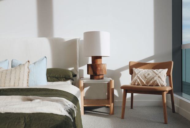 A bright, serene bedroom corner with a partially visible bed dressed in layered white, cream, pale blue and olive-green bedding. A timber bedside table supports a sculptural timber lamp with a white cylindrical shade, while a mid-century-style timber armchair with a textured cream cushion sits beside a large window. The room has white walls, light carpet and strong natural sunlight.