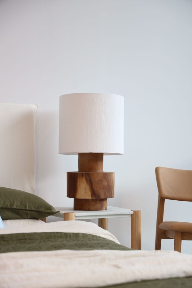 A softly lit bedroom vignette featuring a neatly made bed with olive-green and cream bedding, a sculptural wooden table lamp with a white fabric shade on a small light-wood bedside table, and a simple wooden chair against a pale wall. The composition has a calm, natural, minimalist feel.