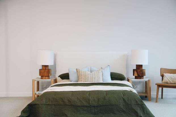 A calm, simply styled bedroom viewed straight on, featuring a centrally positioned bed with a white upholstered headboard, layered white and pale blue pillows, and a dark olive-green quilt. Matching timber bedside tables hold paired white lamps with chunky wooden bases. A tan occasional chair sits to the right against white walls, with pale carpet flooring throughout.