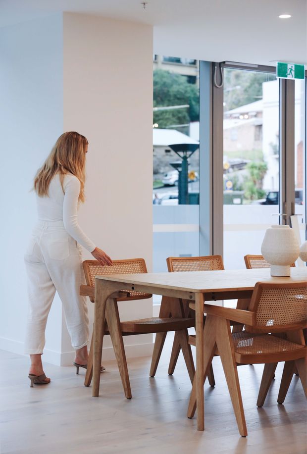 Bright contemporary dining area with a light timber dining table and matching cane-back chairs, set against white walls and pale timber flooring. Large glazed doors or windows provide views to the exterior, and a sculptural ceramic table lamp sits on the dining table. A person is standing beside the table.