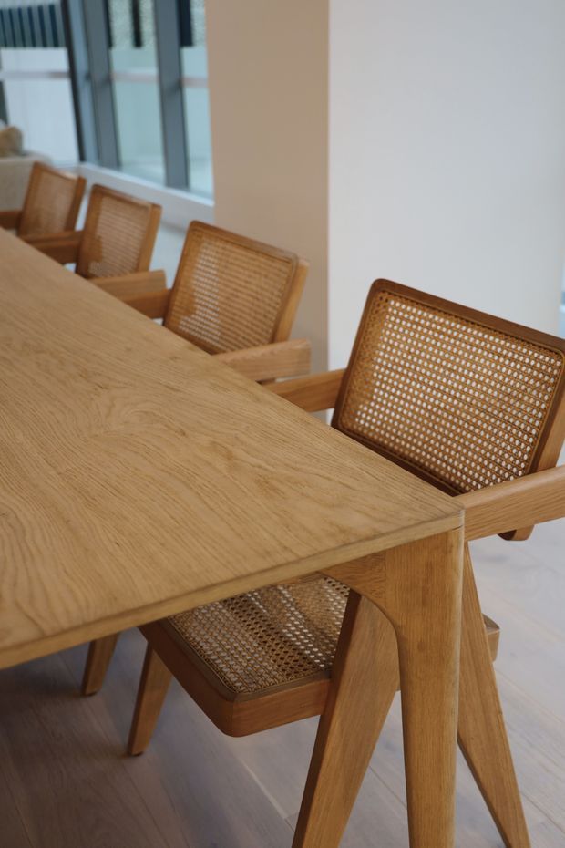 A warm, minimalist dining area featuring a large light-oak dining table with substantial angled legs and matching wooden dining chairs with woven cane backs and seats. Tall dark-framed windows line the left wall, while pale walls and light timber flooring create a bright, natural interior.