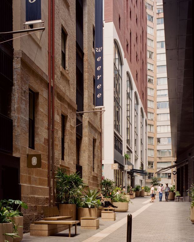 A narrow urban pedestrian laneway framed by historic sandstone masonry on the left and a newer red-brick and rendered building on the right. Tall arched windows, vertical banners, planted raised planters, built-in sandstone seating, paving, café-style furniture, and surrounding multi-storey buildings create a refined commercial streetscape.
