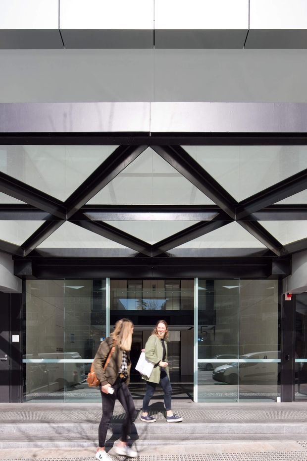 Street-level view of a contemporary commercial building entrance. A glazed entry is framed by dark metal structure and a prominent geometric triangular and diamond-patterned canopy or facade, with broad overhead glazing and polished stone or concrete surrounds. People are walking in front of the entrance, and cars are visible only as reflections through the glass.