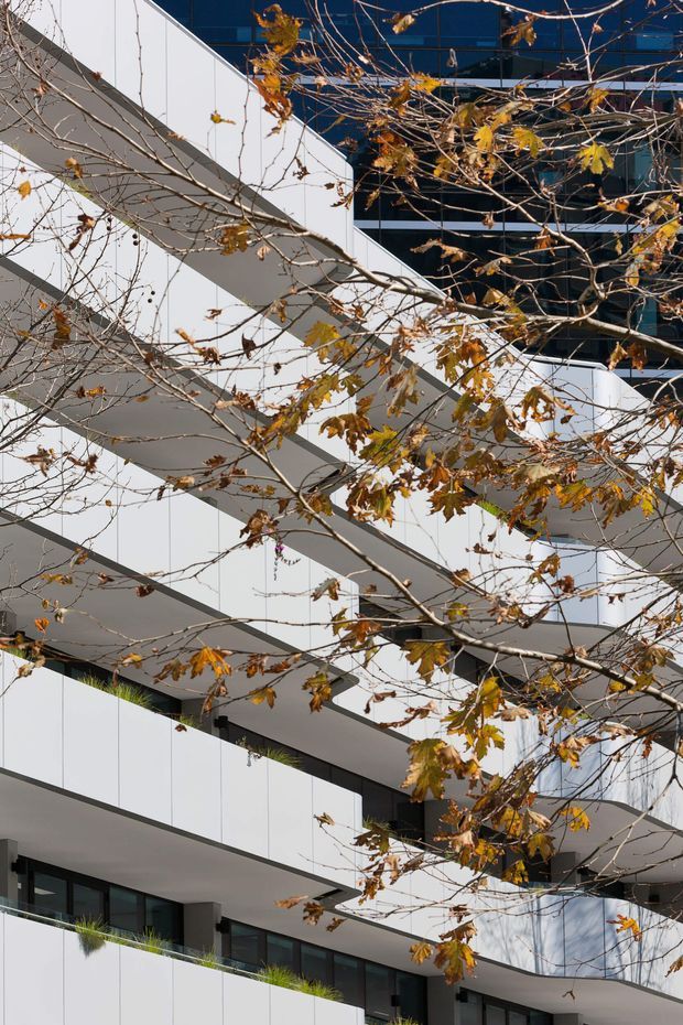 Exterior architectural photograph of a contemporary multi-storey building with stacked, projecting white balcony slabs and dark floor-to-ceiling glazing. Autumn tree branches partially frame the facade, with planted balcony edges visible near the lower levels.