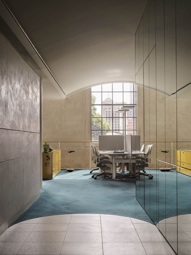 A refined open-plan office interior viewed from a tiled entry area. The space features a central cluster of contemporary workstations and ergonomic office chairs, vivid blue carpet, bright yellow built-in storage, reflective full-height glass or mirrored cabinetry along the right wall, textured grey wall panels on the left, and a large gridded window overlooking an urban setting. The curved ceiling and clean geometric lines create a polished, modern commercial workplace atmosphere.
