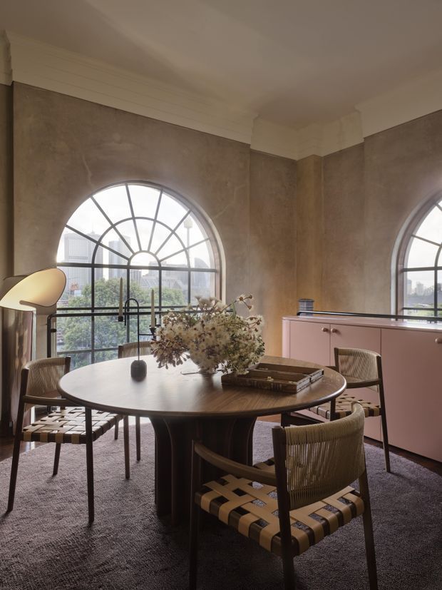 A refined dining room with a round dark-wood pedestal dining table surrounded by woven-seat dining chairs. Large arched steel-framed windows dominate the room, providing abundant natural light and city views. The space features warm taupe textured walls, white decorative ceiling panelling and cornices, a muted pink built-in sideboard, a floor lamp, and a dark textured carpet. The overall atmosphere is sophisticated, warm, and contemporary with classic architectural detailing.
