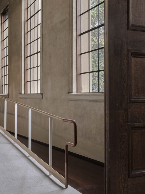 Interior view of a refined circulation space or landing with tall multi-pane steel-framed windows, aged plaster walls, dark timber flooring, and a white railing with a distinctive copper-toned handrail. A large dark timber panelled door occupies the right side of the frame, contributing to a traditional, heritage-inspired character.