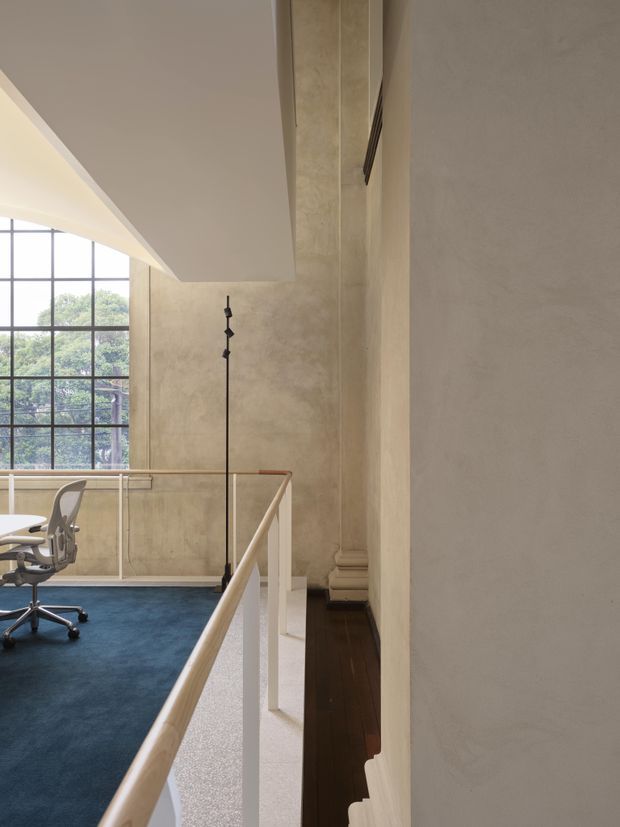 Interior view of an elevated open-plan workspace or landing, featuring a large black-grid window, blue carpet, white office chair, pale timber and white balustrades, dark timber flooring, and tall textured plaster or stone-finished walls with classical moulded detailing. The space has abundant natural light and a refined, quiet atmosphere.