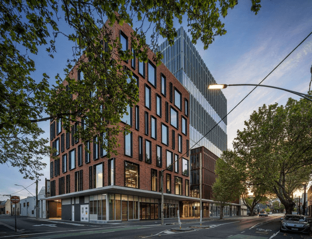 Street-level exterior view of a contemporary multi-storey urban commercial building. The foreground structure combines a red brick facade with repetitive narrow black-framed windows and a glazed ground-floor frontage, while a taller adjoining tower is clad in vertical grey metal fins. Mature street trees, overhead street lighting, sidewalks and roadway define the urban setting.