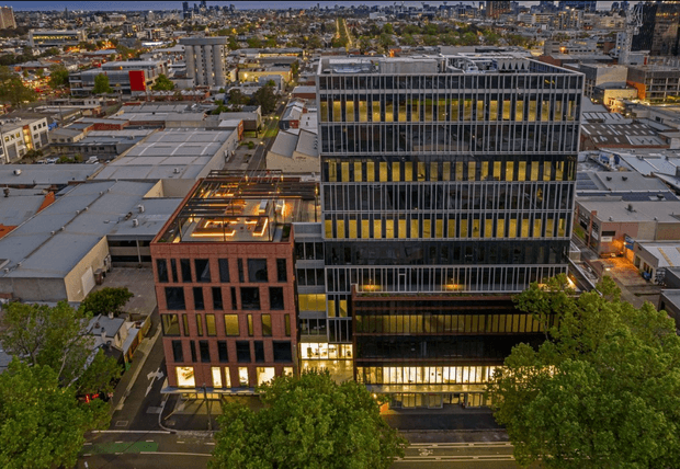 Aerial dusk view of a dense urban commercial precinct, dominated by a contemporary multi-storey office building with a dark metal-and-glass facade, strong vertical fins, and warm illuminated windows. A smaller adjacent brick office building has a rooftop terrace or courtyard with linear lighting. Mature street trees, surrounding low-rise commercial buildings, and a distant city skyline are visible.