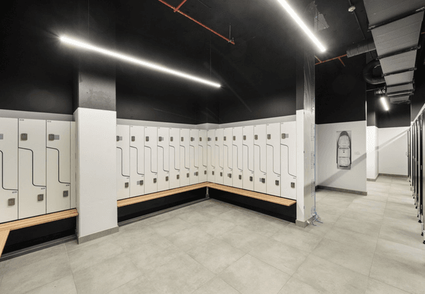 A modern commercial locker room or changing facility with extensive white built-in lockers arranged along two walls, integrated timber bench seating, a large pale tiled floor, and exposed black-painted ceiling services. Linear LED strip lighting and visible ductwork create an industrial contemporary atmosphere.