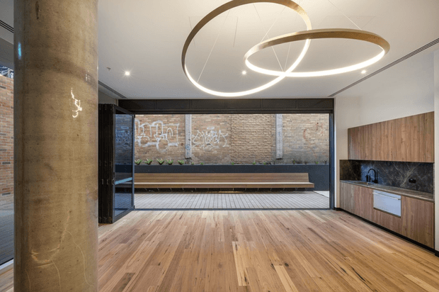 A refined open-plan interior with a warm timber floor, extensive operable glazing opening to a brick-lined courtyard, and a compact timber and dark stone kitchen along the right wall. A large cylindrical concrete column, exposed brickwork, black-framed glazing, and sculptural circular pendant lights create a polished industrial-contemporary character.