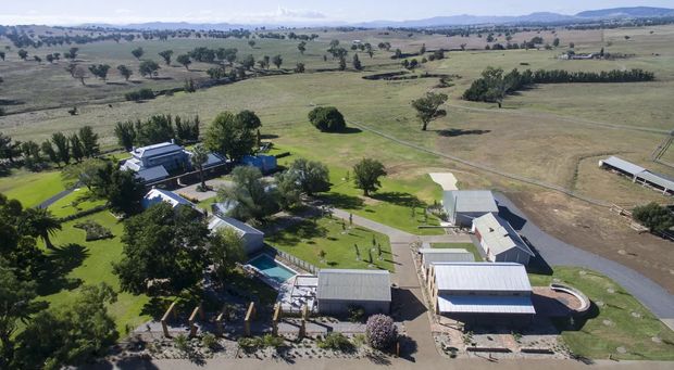 Aerial view of a rural property and homestead set within expansive open farmland. The compound includes several low-rise buildings, long shed-like structures, tree-lined gardens, paved access paths, and a visible swimming pool, with rolling agricultural landscape extending into the distance.