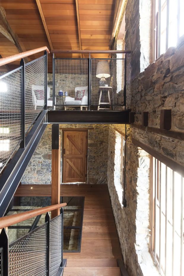 A multi-level interior circulation space in a converted stone building, featuring exposed rough-stone walls, warm timber ceilings and floors, timber doors, and industrial-style black metal stairs and mezzanine balustrades infilled with wire mesh. Large windows bring in bright natural light, while a small seating/work area is visible on the upper landing.