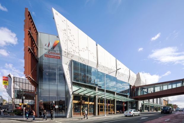 Exterior street-level view of a contemporary multi-storey commercial building with a prominent angular white facade, extensive curtain-wall glazing, a glazed entrance canopy, and a pedestrian bridge connecting to an adjoining wing. The building is set in an urban streetscape under a bright blue sky.