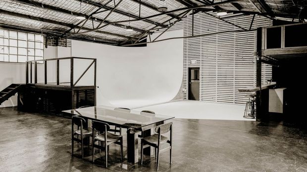 A spacious industrial-style studio or commercial space with a polished concrete floor, exposed structural steel roof framing, reflective insulated ceiling panels, large warehouse glazing, and a white cyclorama backdrop. A long communal table with chairs sits in the foreground, while a raised platform and mezzanine-like structures occupy the sides.