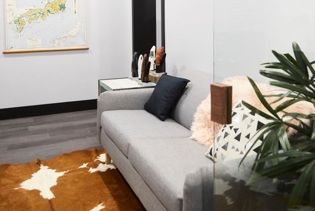 A compact, contemporary living room with a light grey sectional sofa, dark cushions, a cowhide rug, a glass-topped side table, decorative objects, indoor greenery, and a framed map on the wall. The space has dark timber-look flooring, white walls with black trim, and a clean, modern presentation.