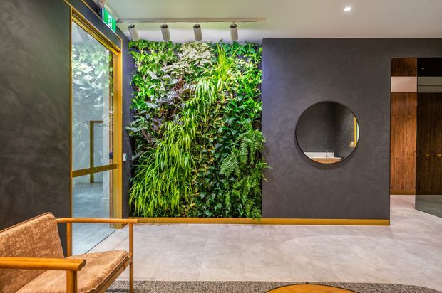 A refined contemporary interior lobby or entry area featuring a dramatic vertical living wall of dense greenery, dark textured wall panels, a round mirror, warm timber and brass-toned detailing, and a glass entry door. A lounge chair and patterned rug sit on a large-format pale grey floor, while built-in timber cabinetry is visible beyond the opening.